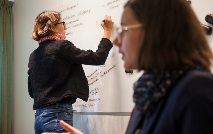 A women is writing on a whiteboard, while another one is talking.